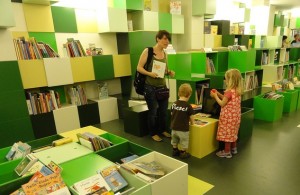 Foto: Regionbibliotek Stockholm. Syntolkning: barnbiblioteksmiljö med mycket gröna inslag i inredningen. En vuxen och två barn är i biblioteket. 