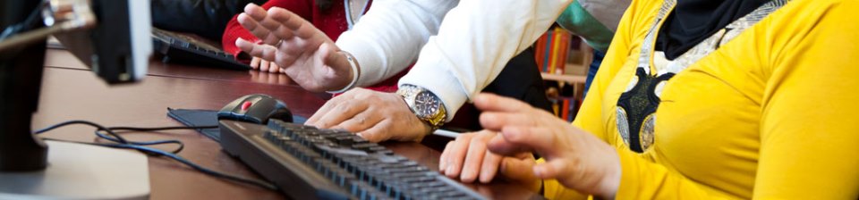 Händer och armar och tangentbord vid dator, närmaste person i klargul kofta, person bortanför i vit tröja och arbandsur, foto