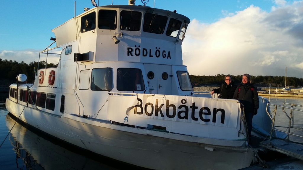 Fartyget Rödlöga syns från sidan med banderoll med texten Bokbåten på relingen och lite skuggor i solljuset