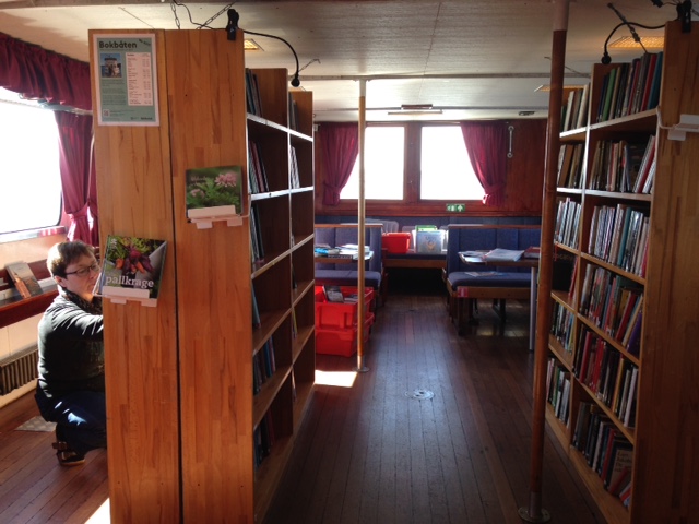Bokbåtens biblioteksrum med hyllor, en person sätter upp böcker