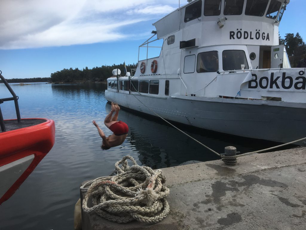 Bokbåtens ena sida vid Ornö och en man i röda badbyxor som slår en volt ner i vattnet